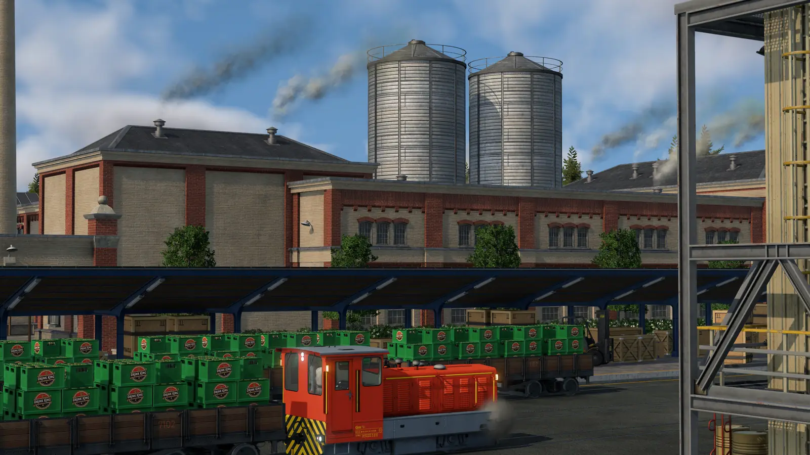 A Transport Fever 3 cargo station: a red shunting engine and a row of wagons loaded with green crates under a long platform roof, with factory buildings and silos behind