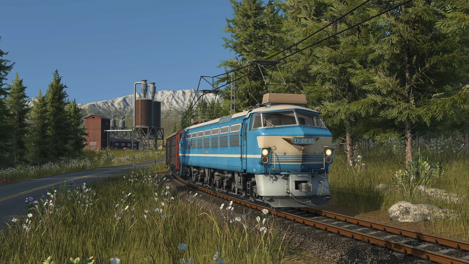 A blue Transport Fever 3 electric locomotive pulling freight wagons under overhead wires, past pine trees, factory silos and a snowy mountain