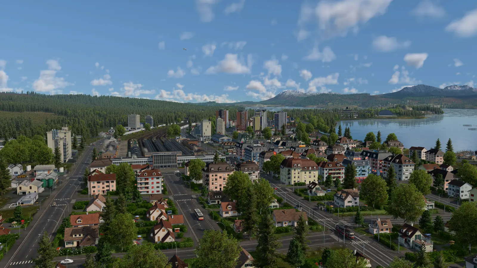 A Transport Fever 3 town of houses and apartment blocks beside a lake, with forested hills and mountains behind it