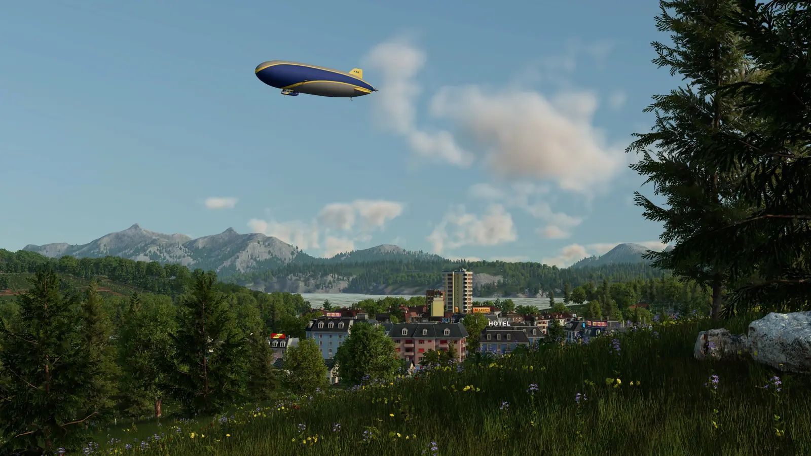 A blue and yellow airship flying over a small Transport Fever 3 town by a lake, with a meadow and pine trees in front and mountains behind