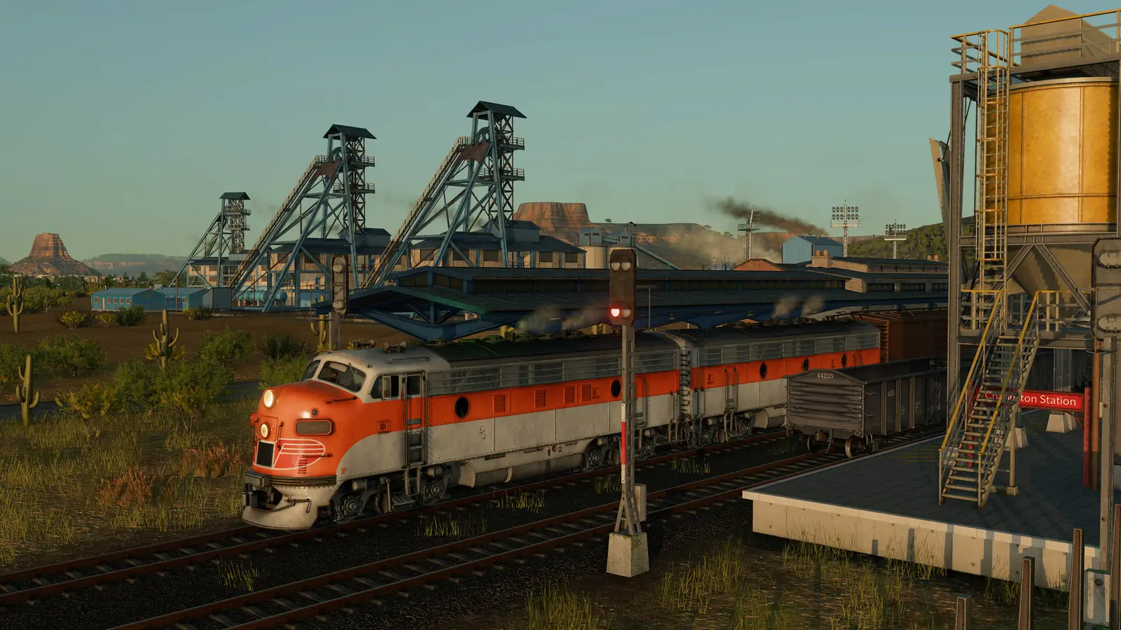 An orange and silver Transport Fever 3 streamlined diesel train waiting at a red signal beside a station platform, with mine headframes and desert mesas behind