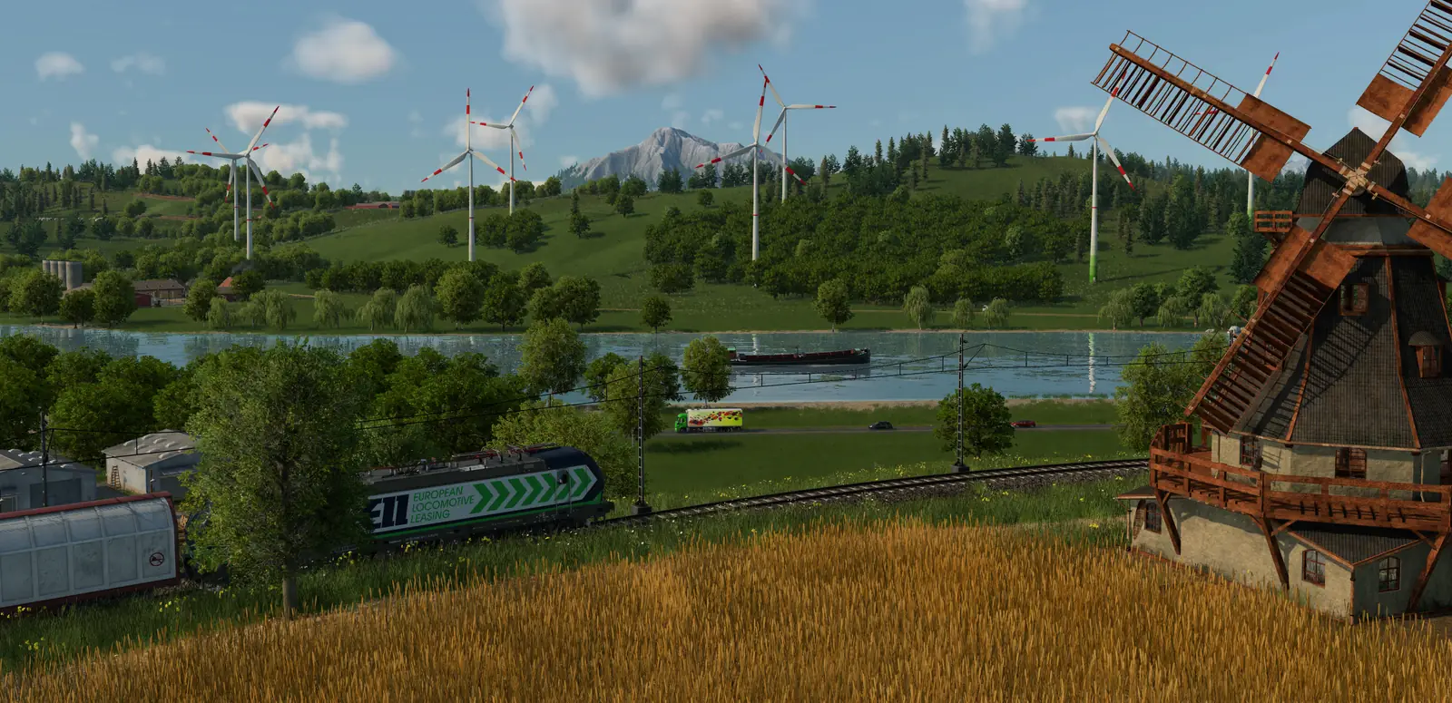 A windmill beside a wheat field in Transport Fever 3, with a freight train on the line below, a barge on the river and wind turbines on the green hills
